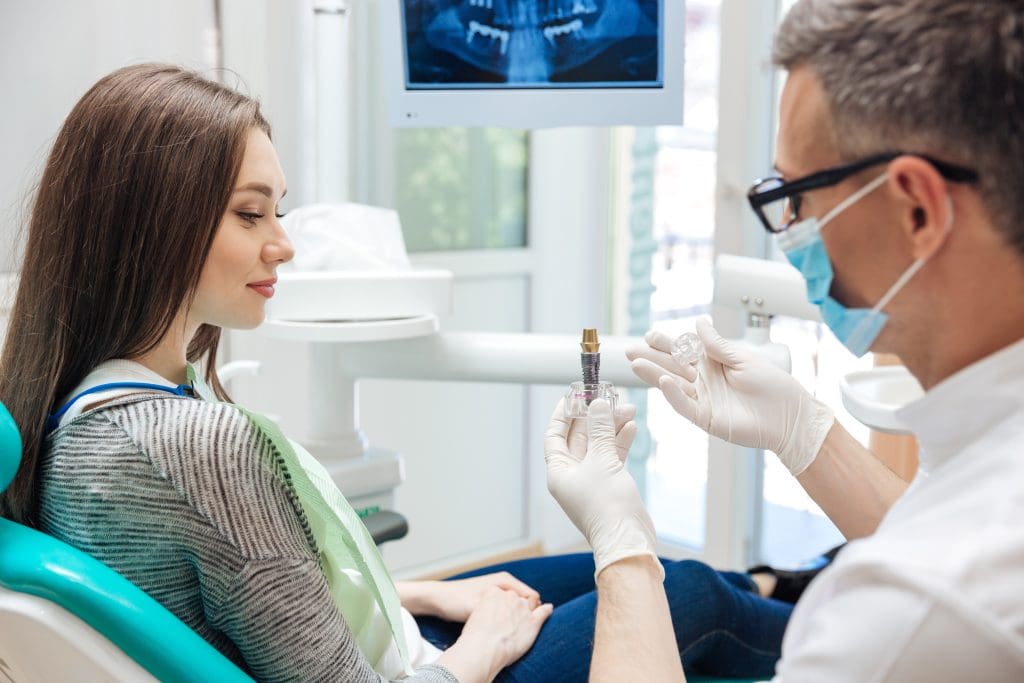 A male dentist, wearing a mask, showing his female patient a dental implant